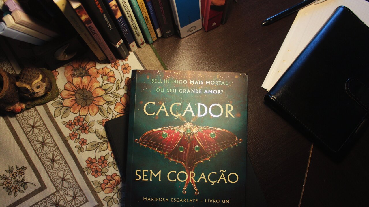 Livro Caçador sem coração, da autora Kristen Ciccarelli, em cima de uma mesa de madeira, com uma toalha florida e cadernos e livros ao redor.