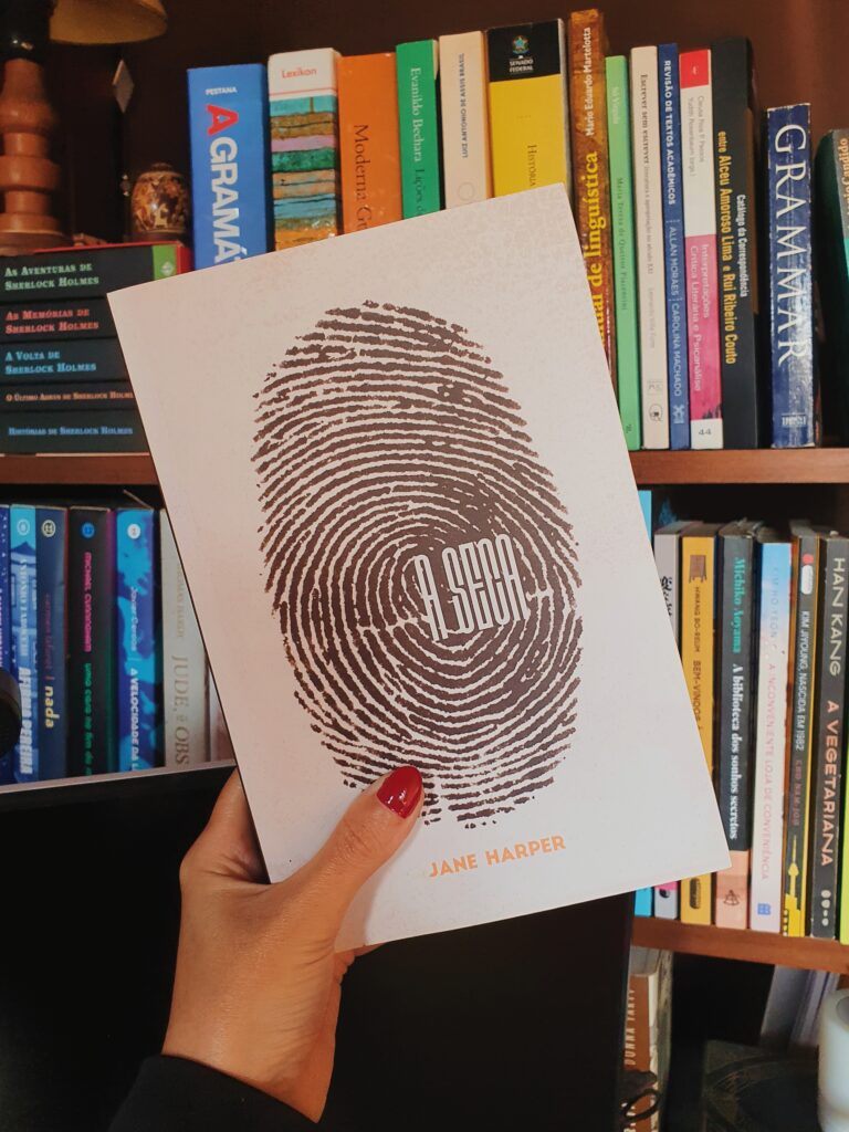 Capa do livro A seca, da autora Jane Harper, sendo segurada em frente a uma estante de livros.