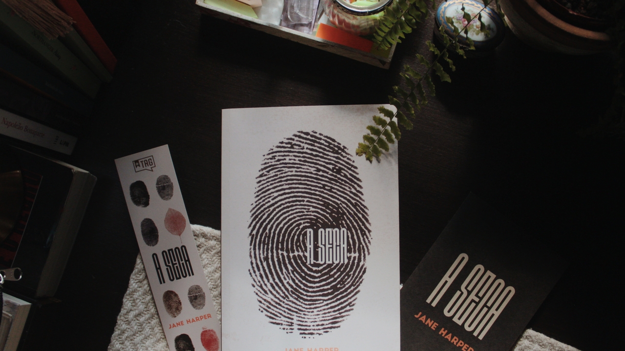 Livro A seca, da autora Jane Harper, em cima de uma mesa, com um marcador de páginas e um guia para o livro, elaborado pela TAG inéditor