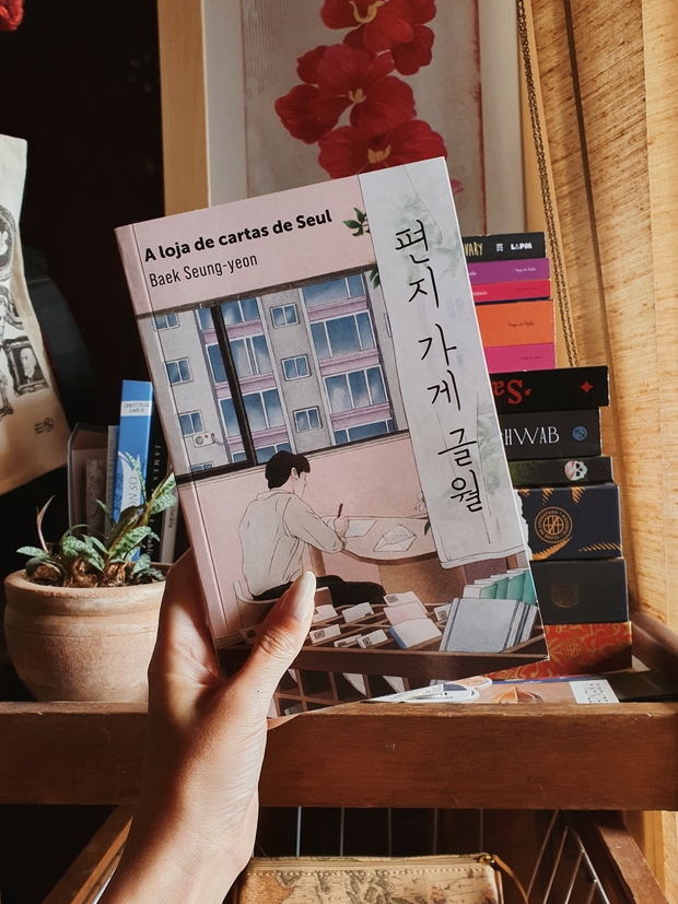 Capa do livro A loja de cartas de Seul, da autora Baek Seung-yeon sendo segurada em frente a um carrinho de livros.